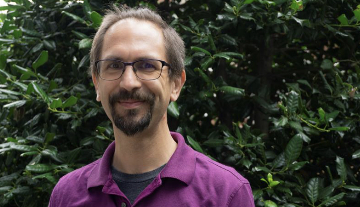 Headshot of J. Swiderski, a white person with brown hair and goatee, wearing a purple polo shirt and standing in front of a large shrubbery or wall of ivy.